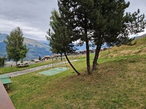 Uma área gramada com mesas de piquenique e baloiços, com vista para uma paisagem montanhosa e edifícios ao fundo.