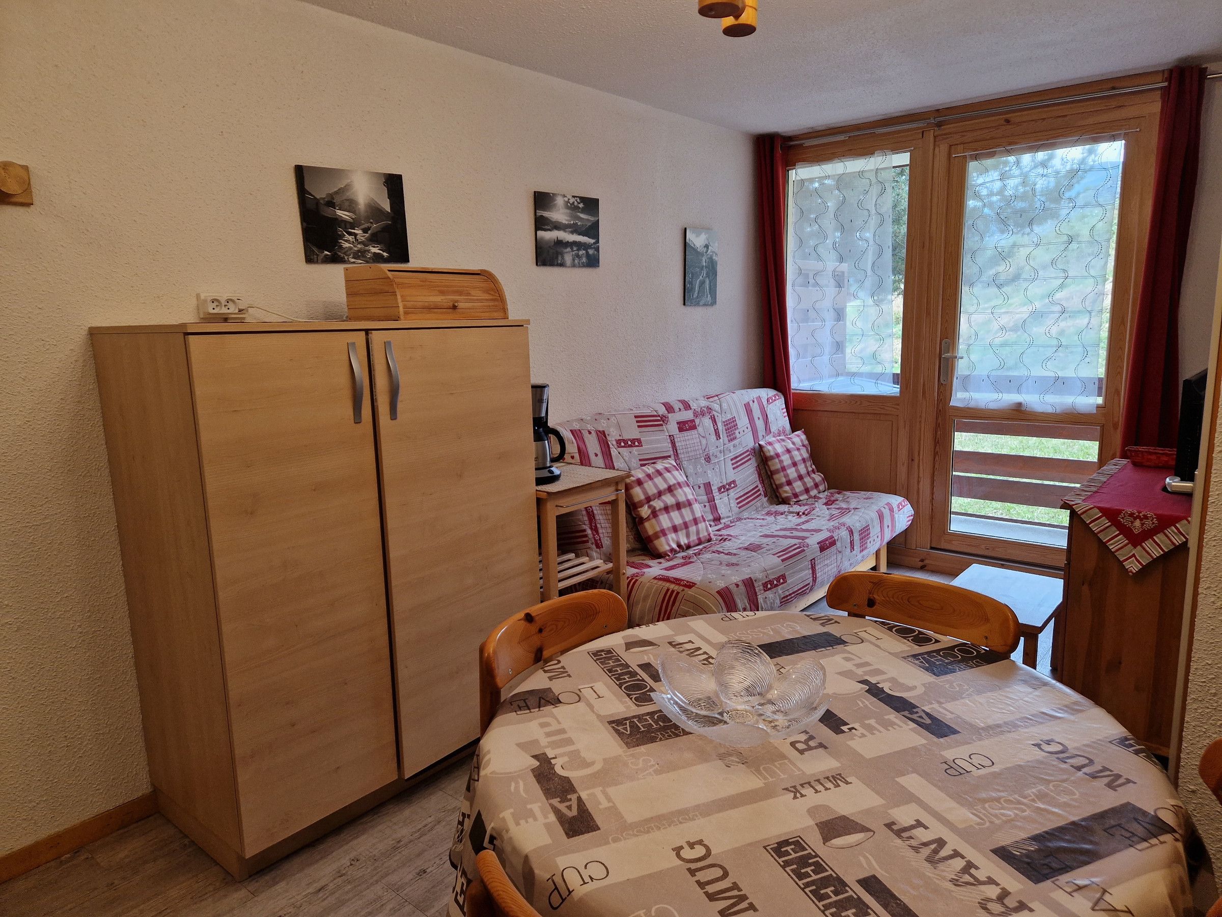 A cozy room with a wooden wardrobe, a patterned sofa, a small table with a coffee maker, and a window with curtains.