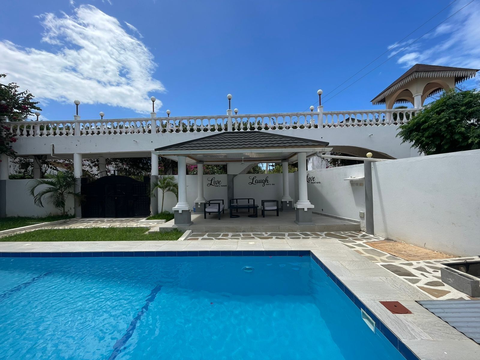 A pool area with a covered seating area, a white fence, and a building with a tiled roof.