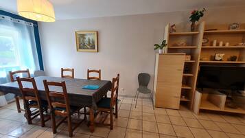 A dining area with a table and chairs, a bookshelf, a painting, and a window with curtains.
