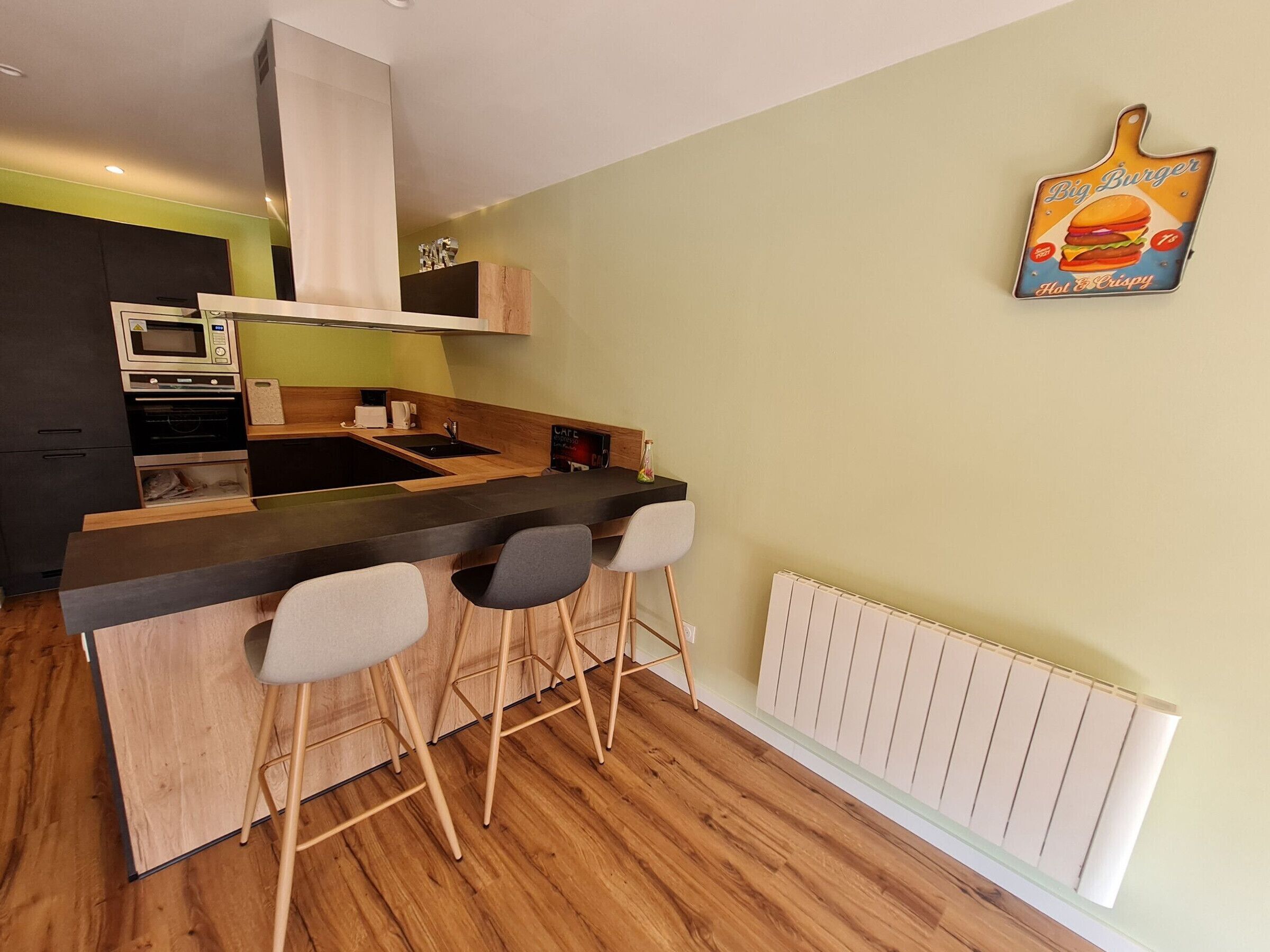 Une cuisine compacte équipée d’un four à micro-ondes, d’un four traditionnel et d’un petit plan de travail. On y trouve trois tabourets de bar et un panneau affiché sur le mur.