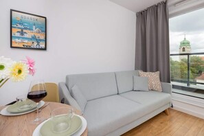 A modern living room with a grey sofa, a dining table with plates and glasses, a vase with flowers, and a cityscape view through a window. - Stylish Apartment with a Balcony | Gdansk Wrzeszcz (Gdansk)