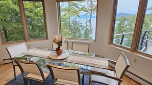 Un coin repas avec une table en verre, quatre chaises et un vase de fleurs. De grandes fenêtres offrent une vue sur les arbres et un lac.