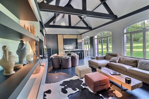 A modern living room with a sofa, ottoman, and coffee table, a kitchen area, and large windows with a view of a green landscape.