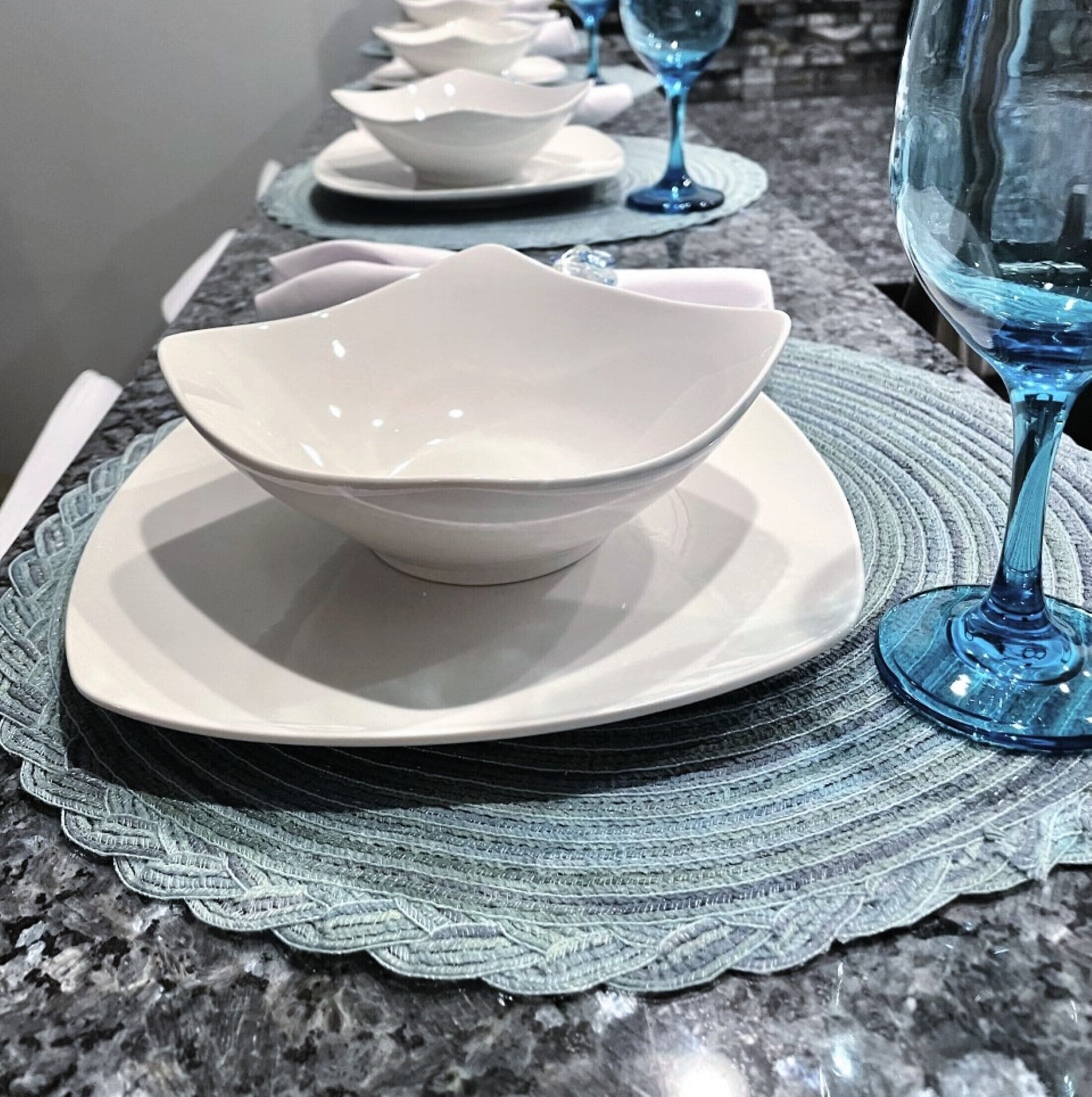 A dining table with white plates and bowls, a blue glass wine glass, and a woven placemat.