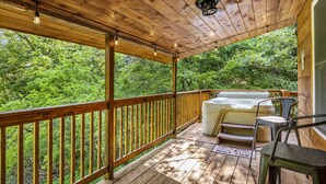 A wooden deck with a hot tub, outdoor seating, and a view of dense greenery.
