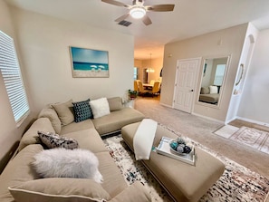 A living room with a sectional sofa, a coffee table with decorative items, a painting on the wall, and a ceiling fan. - Grand Family House With Private Pool Near Disney Parks (Davenport)