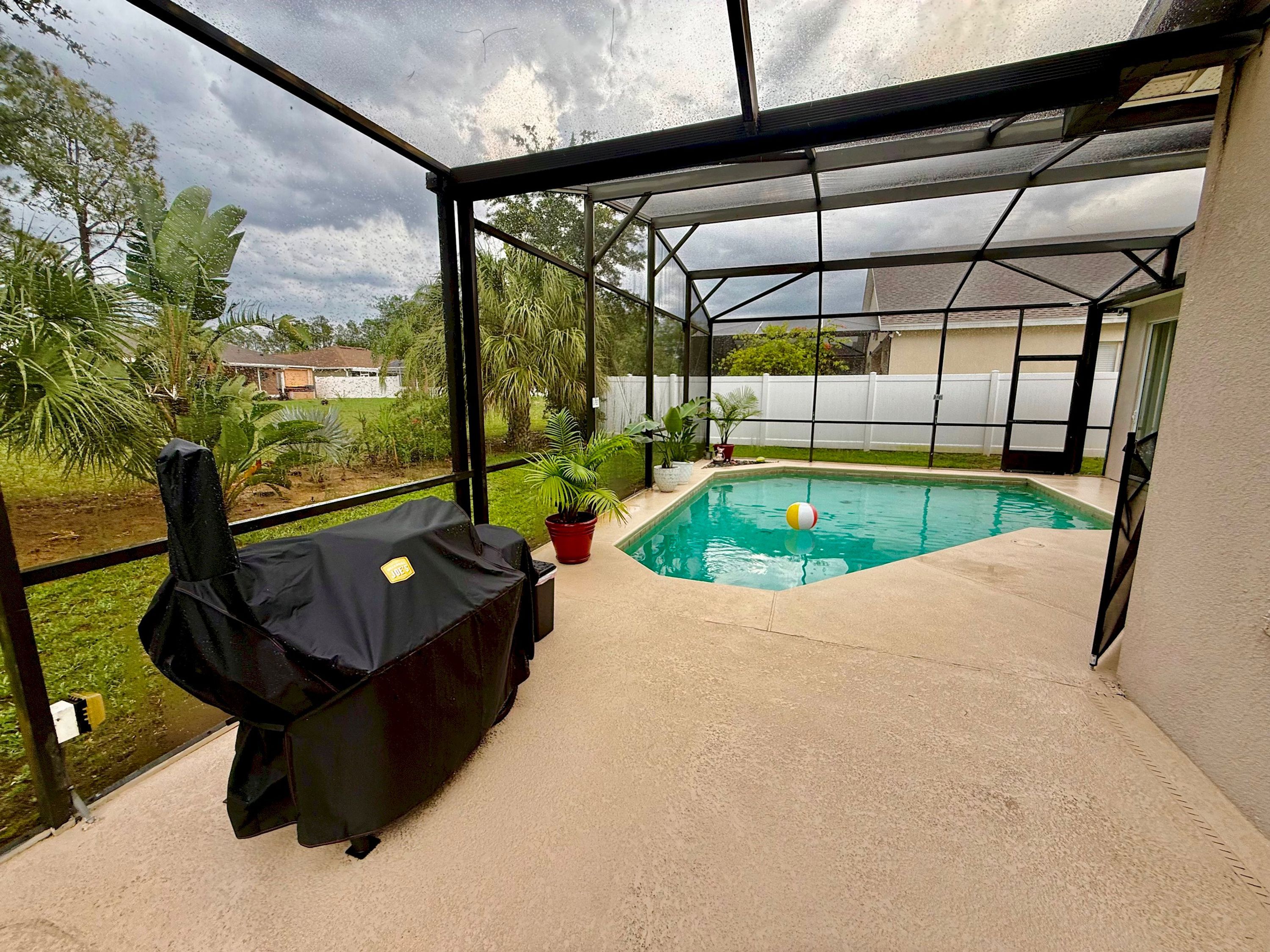 A covered outdoor area with a pool, a grill, and potted plants.