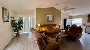 A living room with a brown sofa, a coffee table, and a dining area with a table and chairs. There is a ceiling fan, a potted plant, and a painting on the wall.