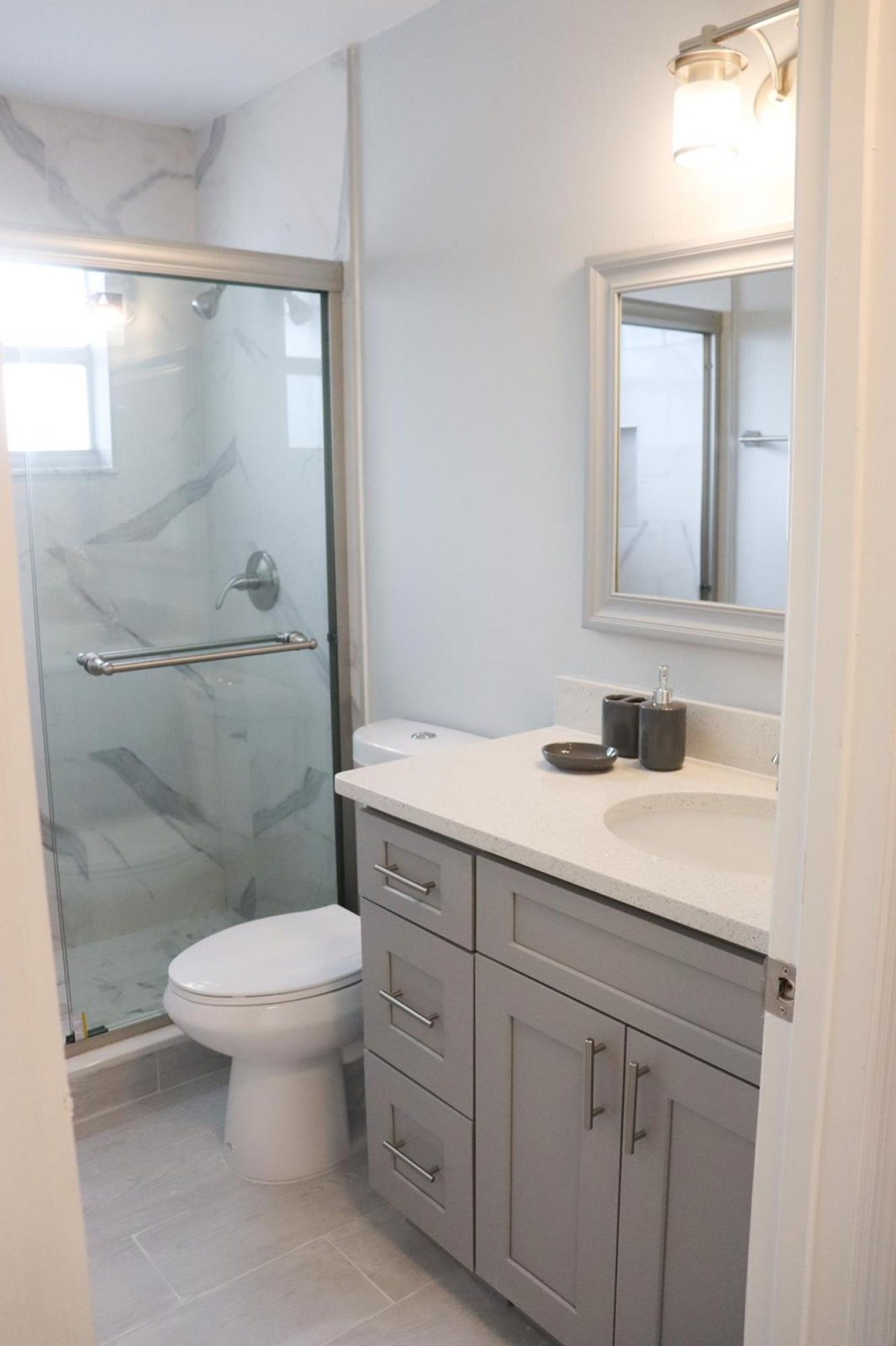 A bathroom with a glass shower enclosure, a toilet, a sink with a mirror above it, and a cabinet with drawers.