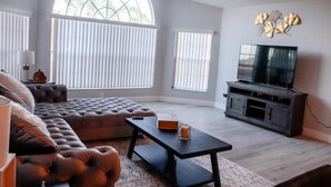 Ein Wohnzimmer mit großem Fenster, Deckenventilator, einer Couch, einem Couchtisch und einem Fernseher auf einem Holzschrank.