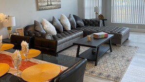 A modern living room with a dark leather sofa, a dining table with yellow placemats, and a large window with blinds. - Grand Executive 3BD Condo Apartment Near Disney Parks 4 (Kissimmee)