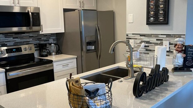 A modern kitchen with white cabinets, stainless steel appliances, a granite countertop, and a coffee menu on the wall. - Grand Executive 3BD Condo Apartment Near Disney Parks 4 (Kissimmee)