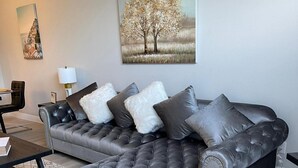 A modern living room with a grey tufted sofa, a coffee table, and a painting of a tree on the wall. - Grand Executive 3BD Condo Apartment Near Disney Parks 4 (Kissimmee)