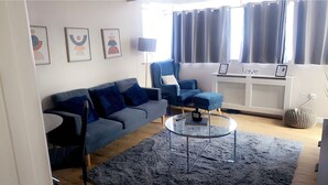 A modern living room with a grey sofa, a blue armchair, a glass coffee table, and a grey rug. There are framed pictures on the wall, a radiator, and a window with curtains. - Remarkable 2-bed Apartment in London (London)