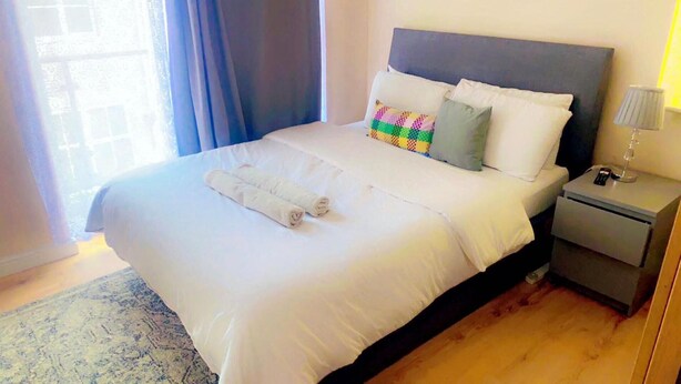 A neatly made bed with white linens and a colorful quilt, a bedside table with a lamp, and a window with curtains. - Remarkable 2-bed Apartment in London (London)