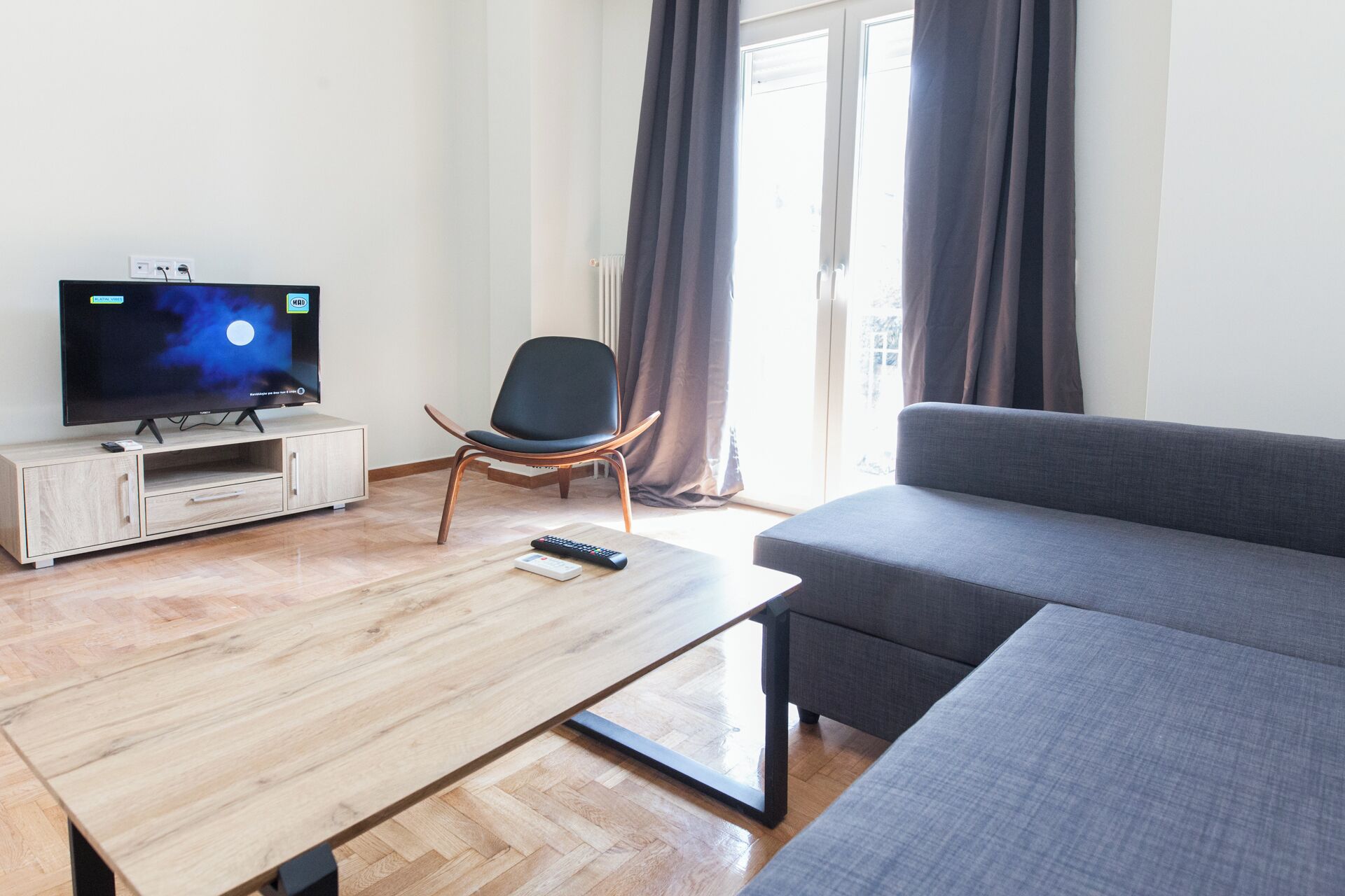 A modern living room with a flat-screen TV, a wooden coffee table, a grey sofa, and a black chair.
