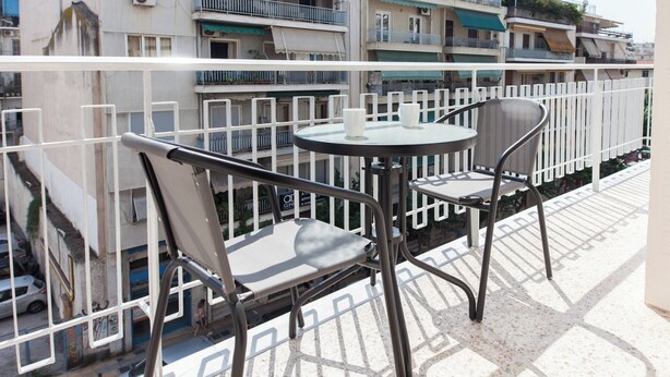 Un balcon avec une table ronde et deux chaises, donnant sur une rue bordée d’immeubles et de voitures garées.