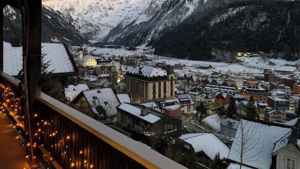 Eine Stadt mit schneebedeckten Bergen im Hintergrund, einem Fluss und Gebäuden mit Schnee auf den Dächern.