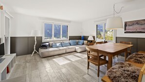 Ein modernes Wohnzimmer mit Sofa, Esstisch und Stühlen. Große Fenster sorgen für natürliches Licht und einen Ausblick ins Freie.