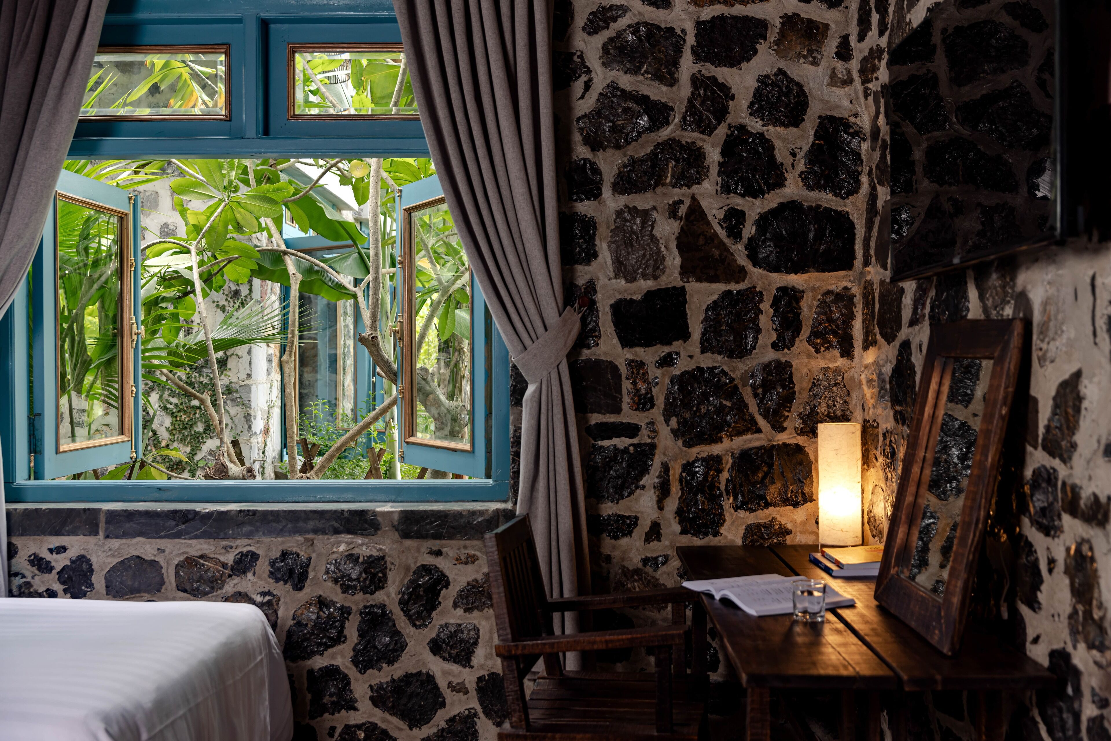 A room with a stone wall, a wooden table with a lamp, a chair, and a bed.