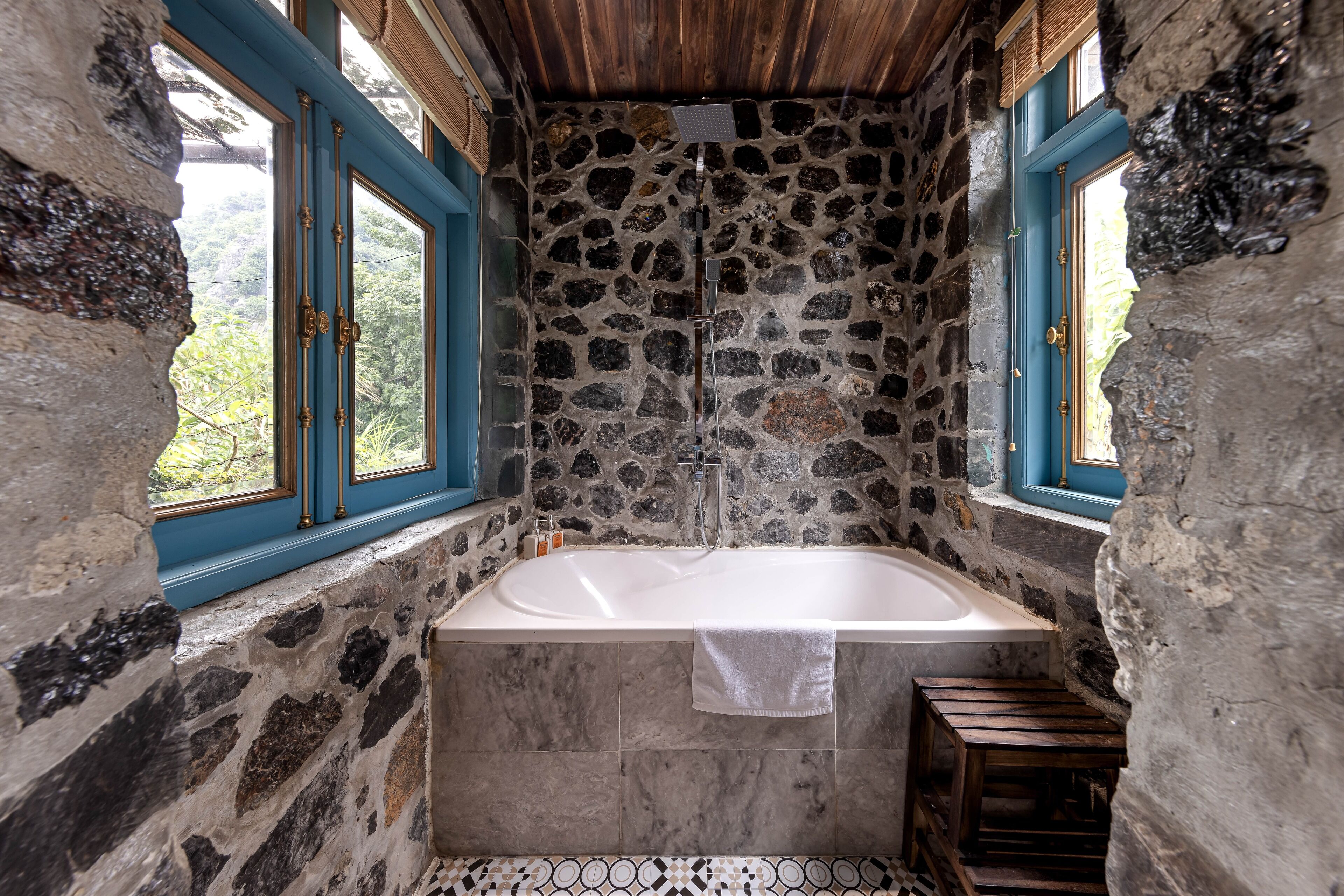 A stone-walled bathroom with a bathtub, a window, and a wooden stool.