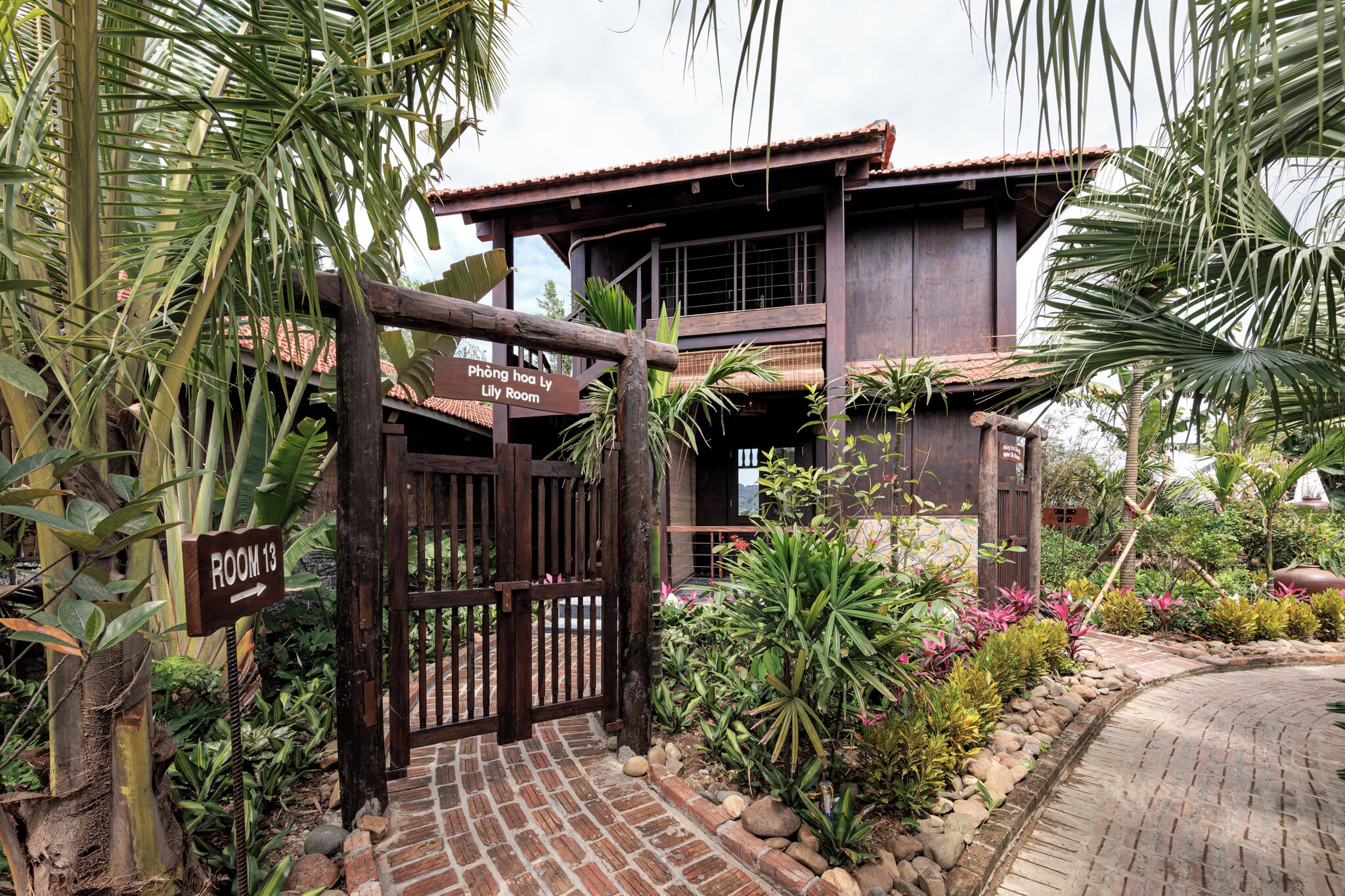 A wooden gate with a sign indicating ROOM 13, a brick pathway, and a tropical garden with various plants.