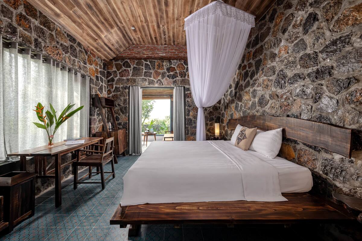 A bedroom with a stone wall, wooden ceiling, a large bed, a desk, and a chair.