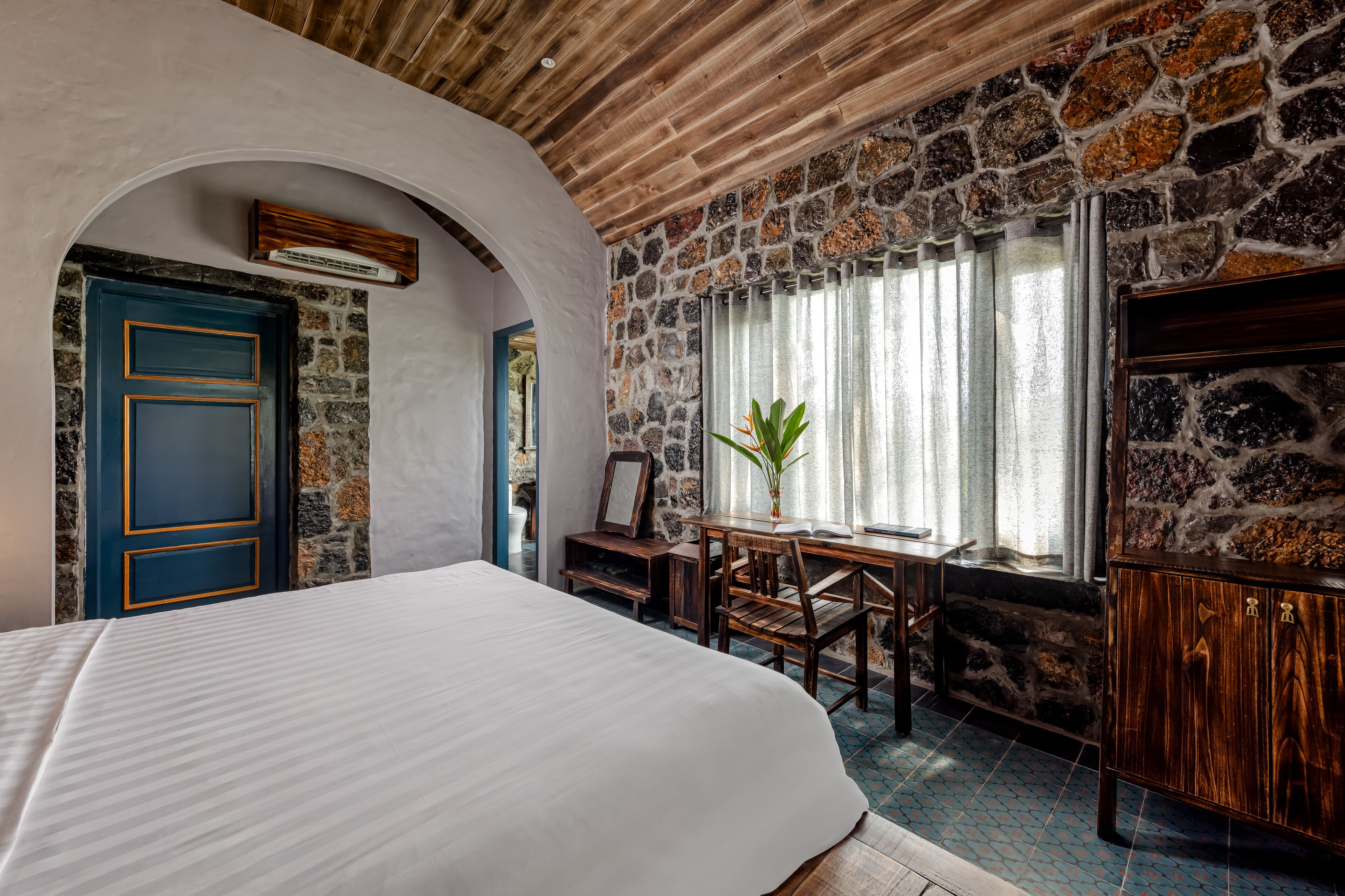 A bedroom with a large bed, stone walls, a wooden ceiling, and a window with curtains.