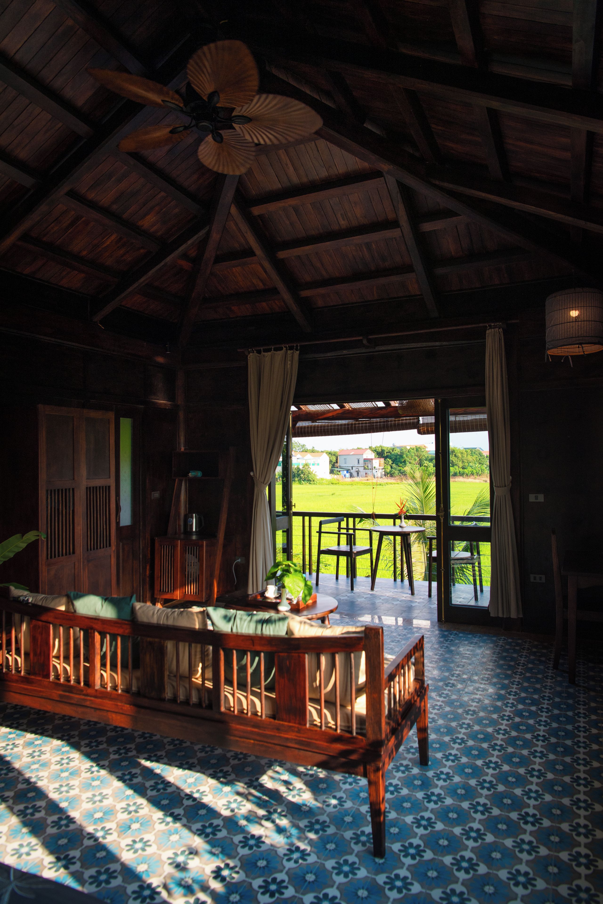 A room with a wooden ceiling, a ceiling fan, a table with chairs, and a view of a green landscape through a doorway.
