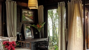 Una habitación con techo de madera, ventilador de techo, mesa de comedor con sillas, sofá, mesita con un florero y un ventanal con cortinas.