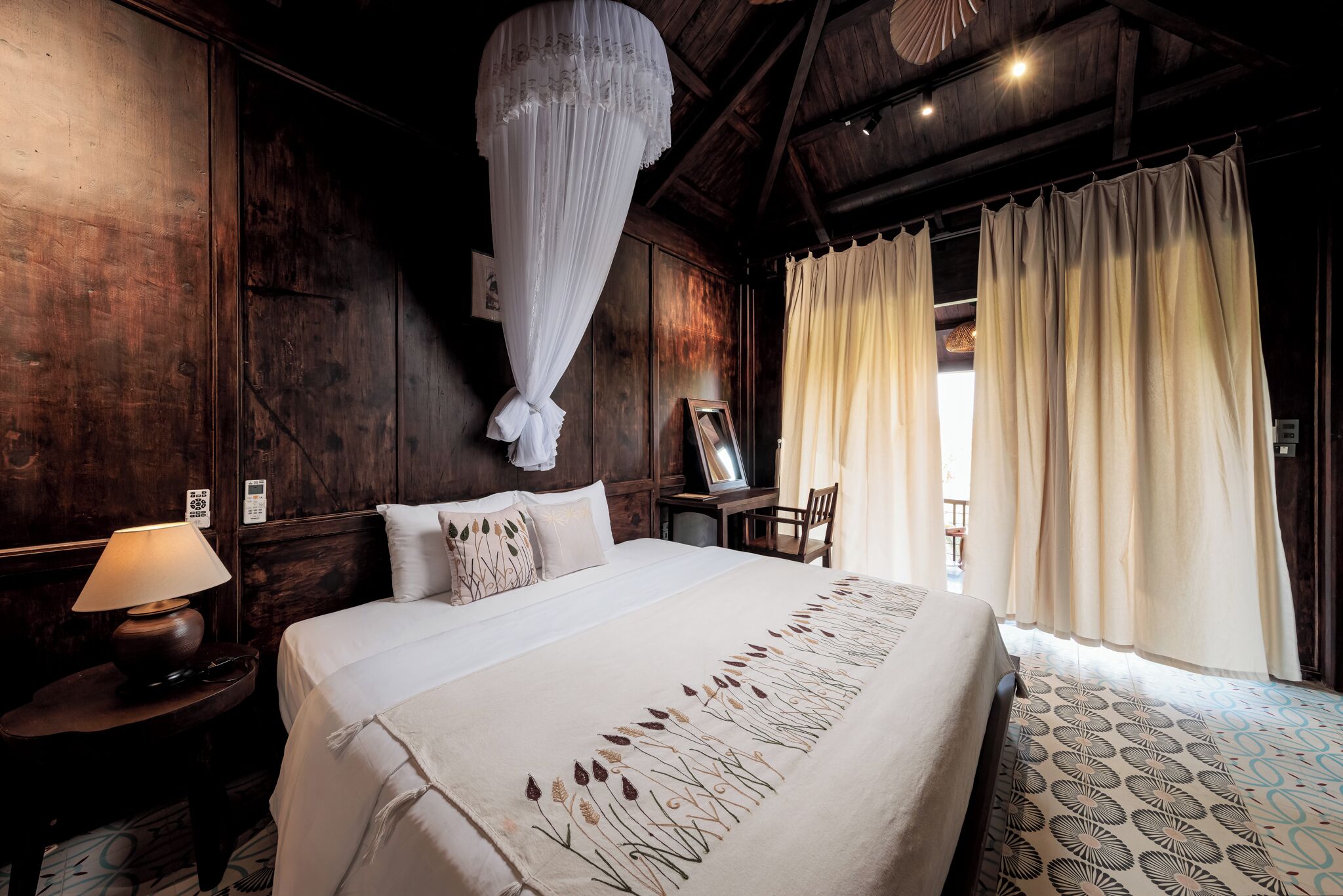 A bedroom with a large bed, a bedside table with a lamp, a wooden headboard, a mirror, and a window with curtains.