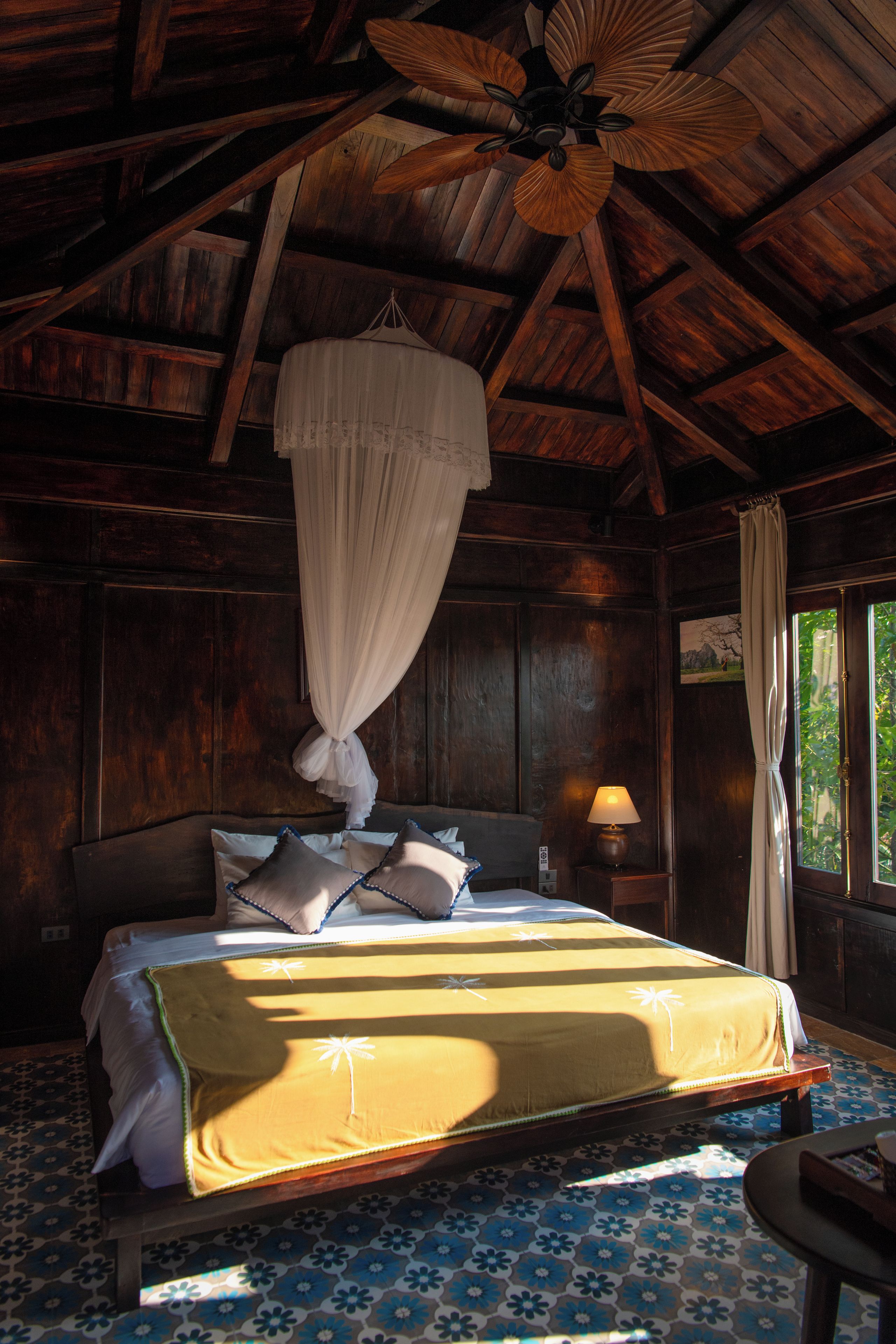 Una habitación con cama, mesita de noche, lámpara, ventana con cortinas y ventilador de techo.