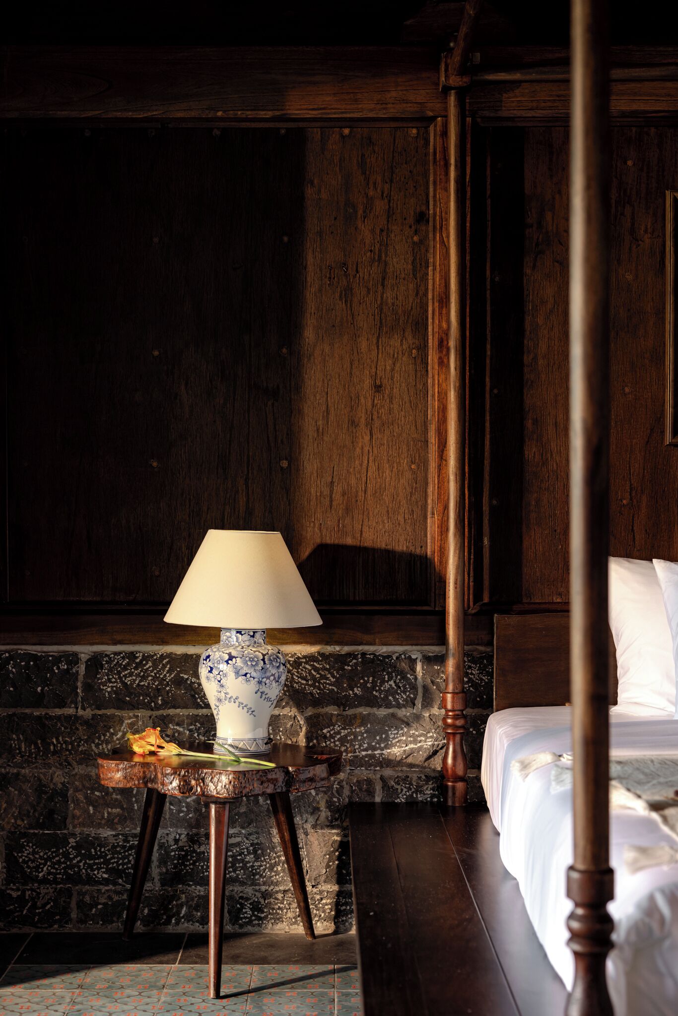 A four-poster bed with a wooden headboard, a bedside table with a lamp, and a stone wall.