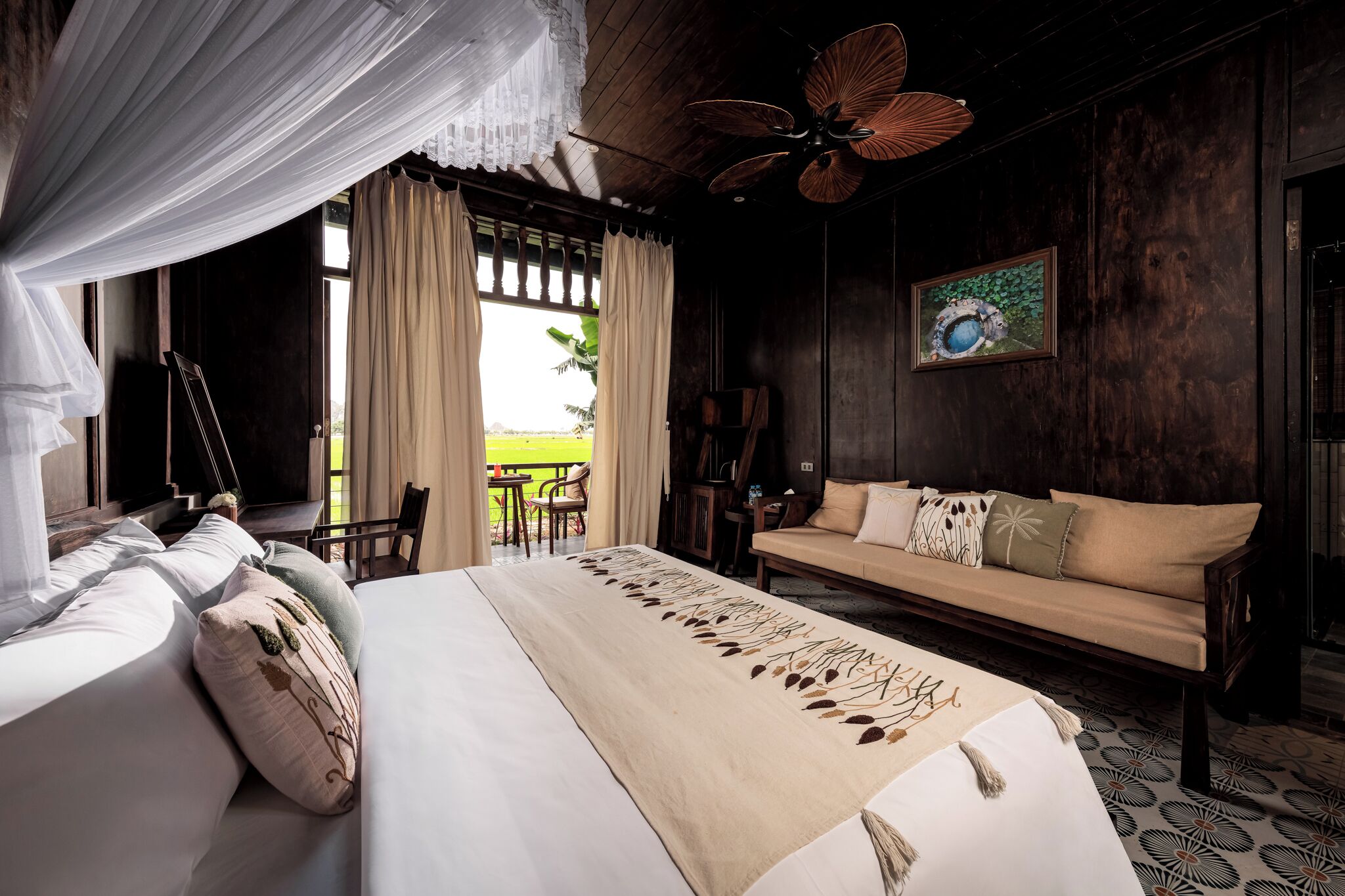 A bedroom with a bed, a sofa, a ceiling fan, a painting, and a view of the outdoors.