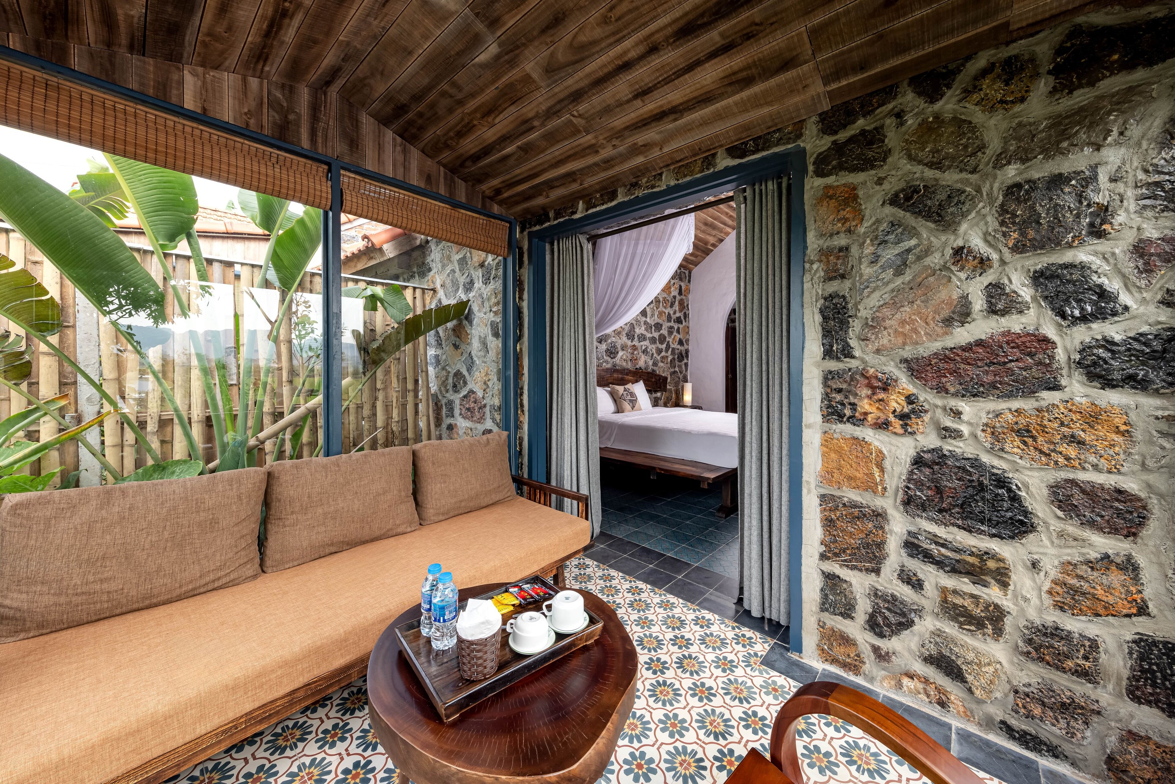 A room with a wooden ceiling, a stone wall, a sofa, a tray with water bottles, and a bed in the background.