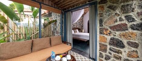 A room with a wooden ceiling, a stone wall, a sofa, a tray with water bottles, and a bed in the background.