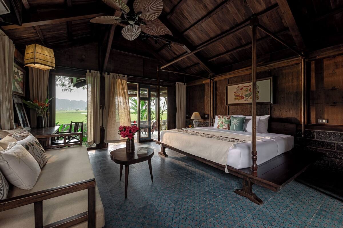 A spacious room with a large bed, a sofa, and a dining table. There is a view of greenery outside the windows.