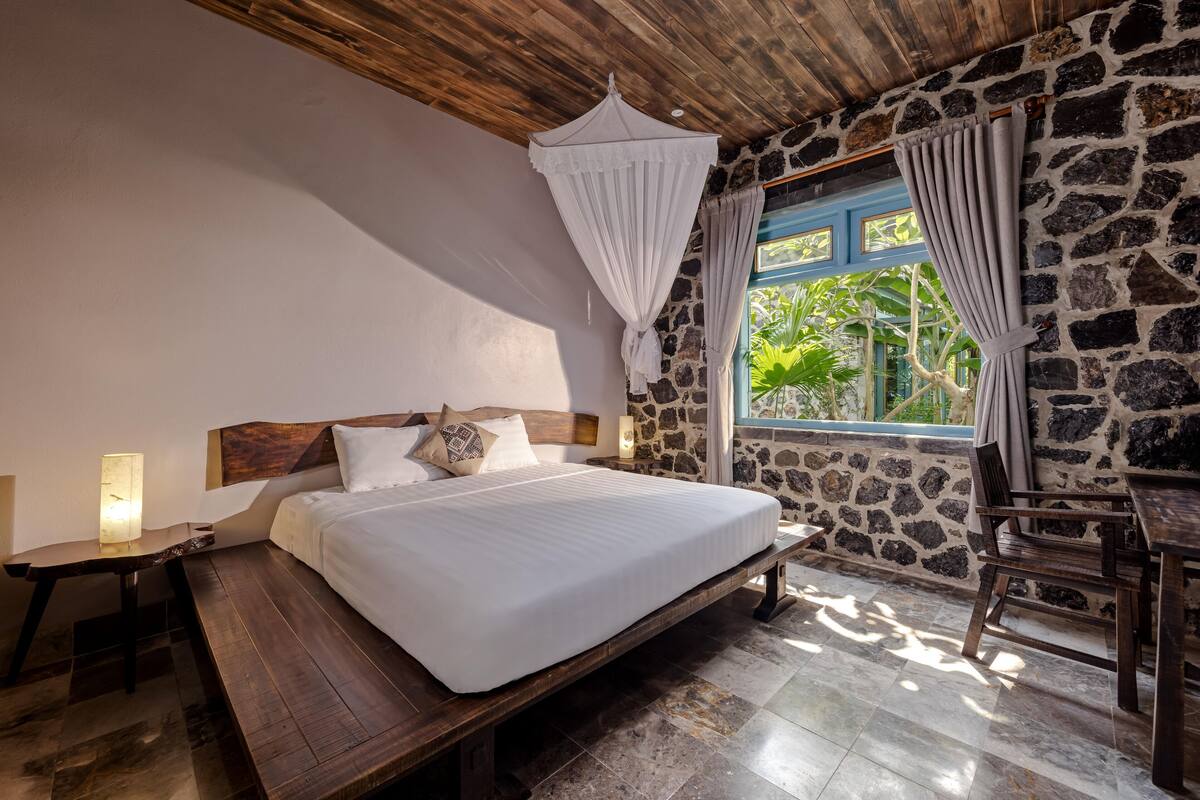 A bedroom with a stone wall, a wooden ceiling, a bed with white bedding, a nightstand, a table, and a window with curtains.
