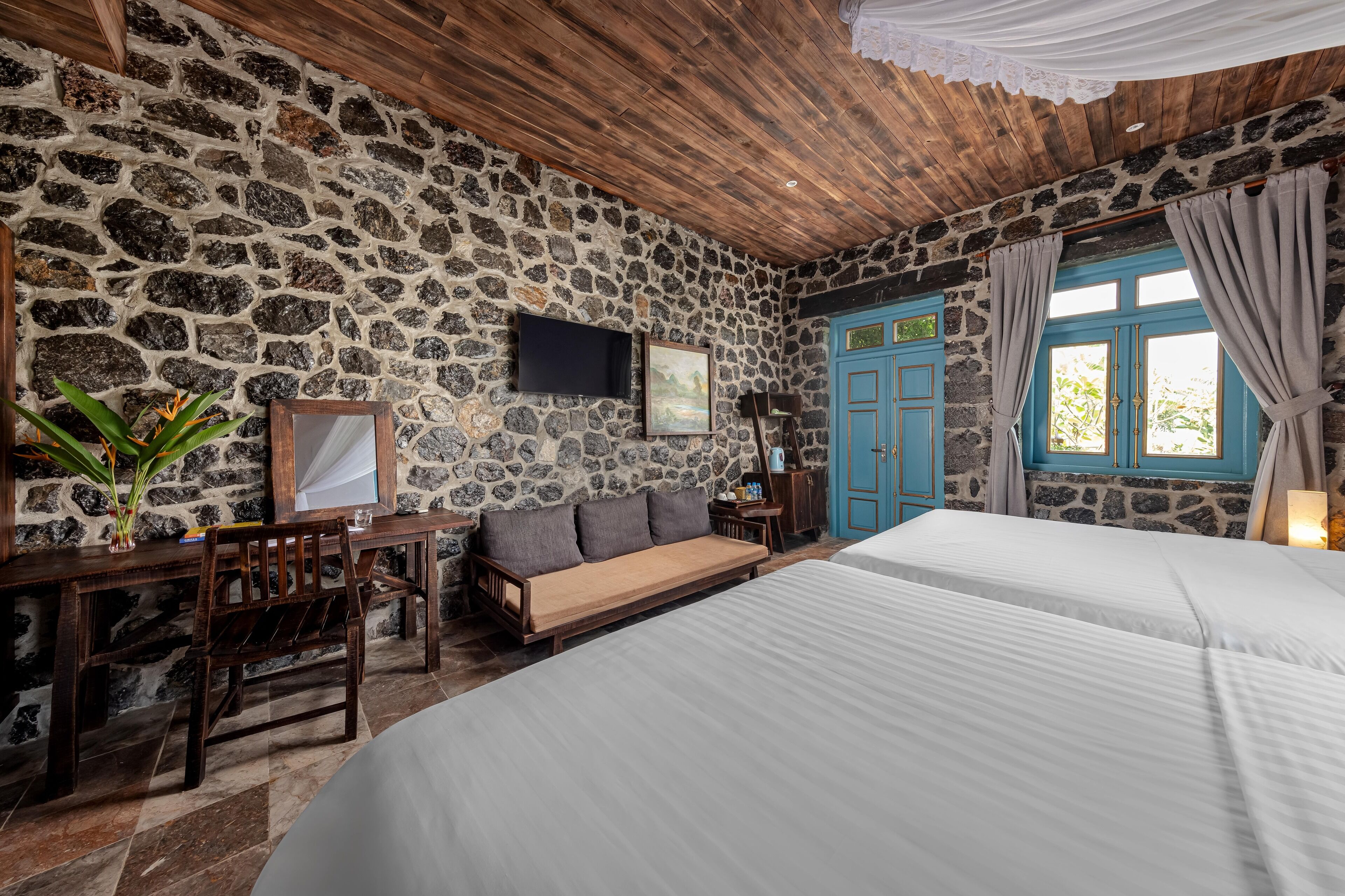 A room with stone walls, wooden ceiling, a bed, a desk, and a chair.