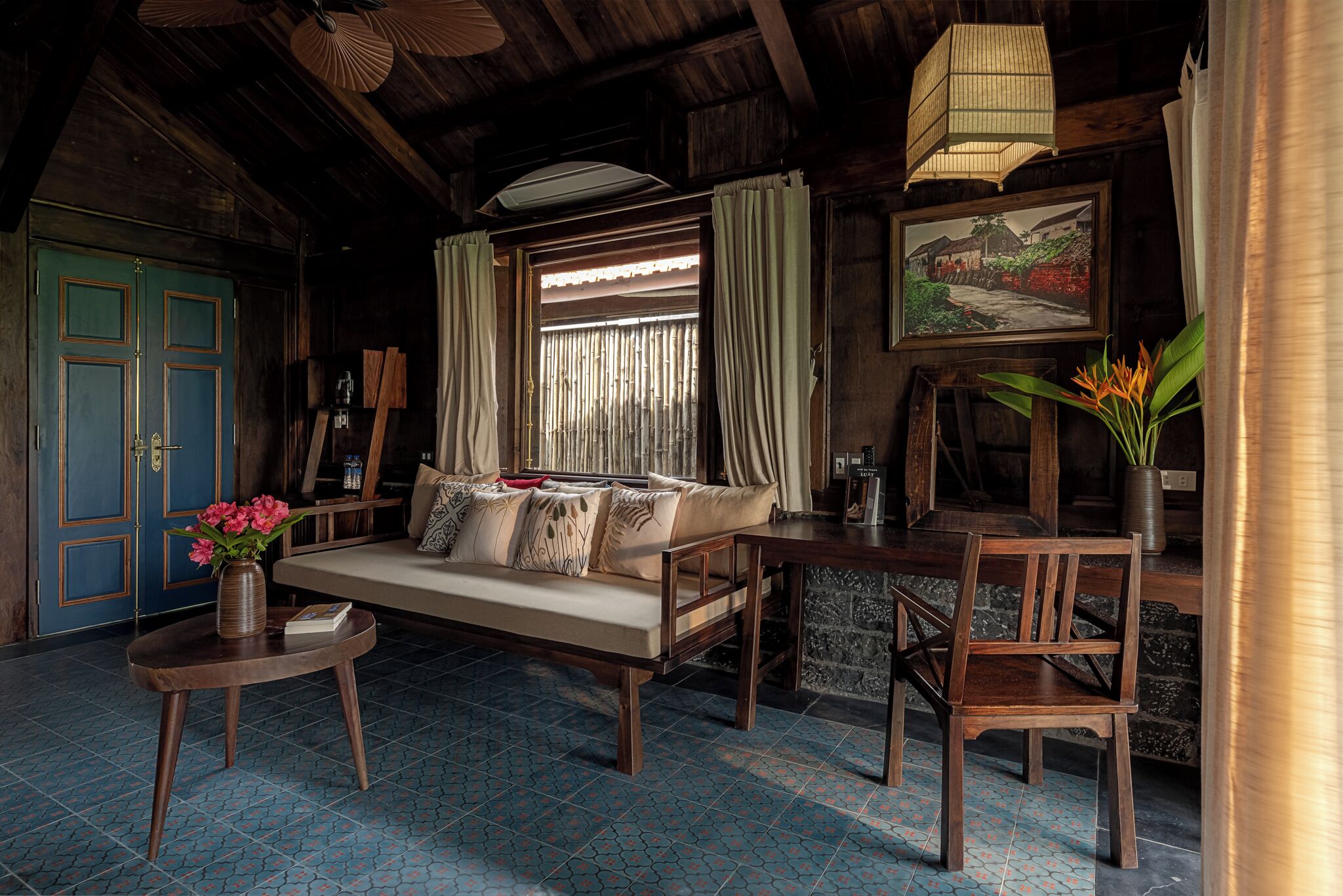 A cozy room with a wooden ceiling, a sofa with cushions, a small round table with a vase of flowers, a chair, and a painting on the wall.
