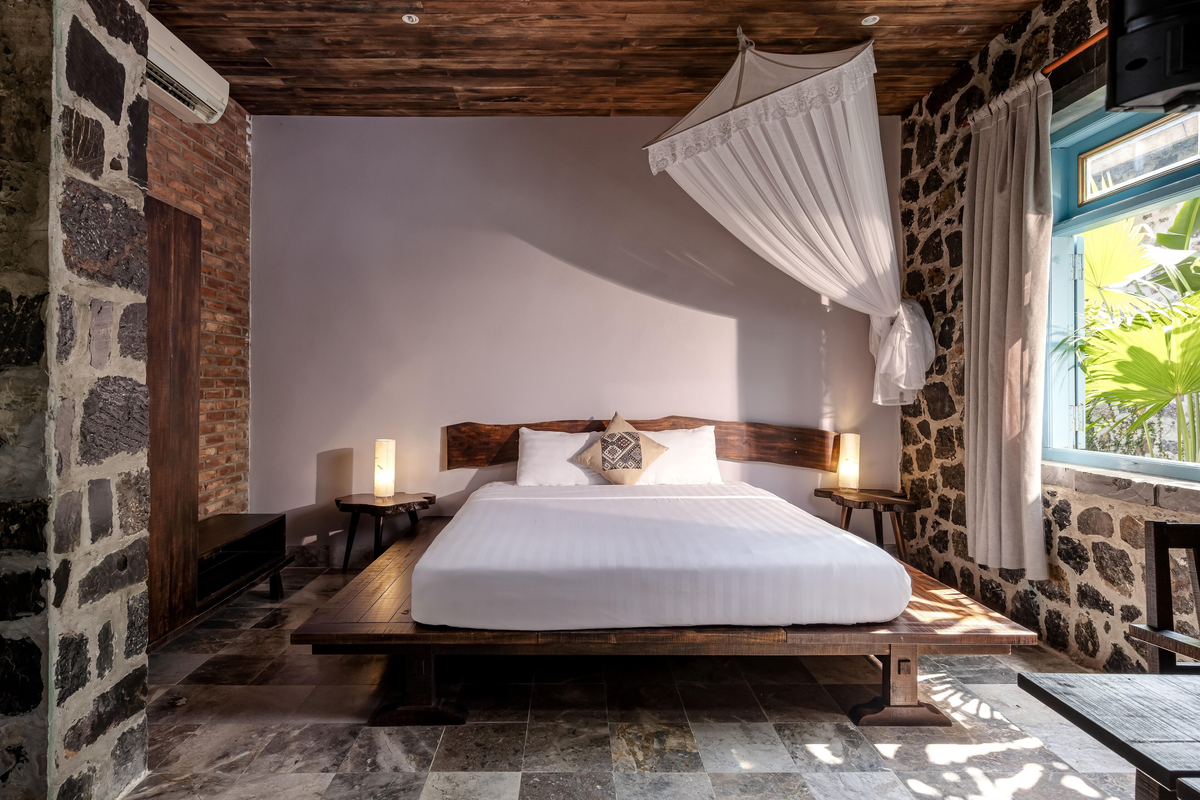 A bedroom with a large bed, stone walls, wooden ceiling, and a window with a view of greenery.