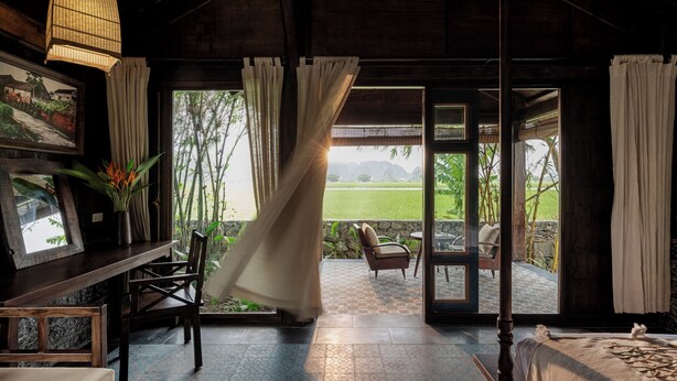 Una habitación con techo de madera, una cama, una mesa con sillas y vista a un jardín a través de un ventanal.
