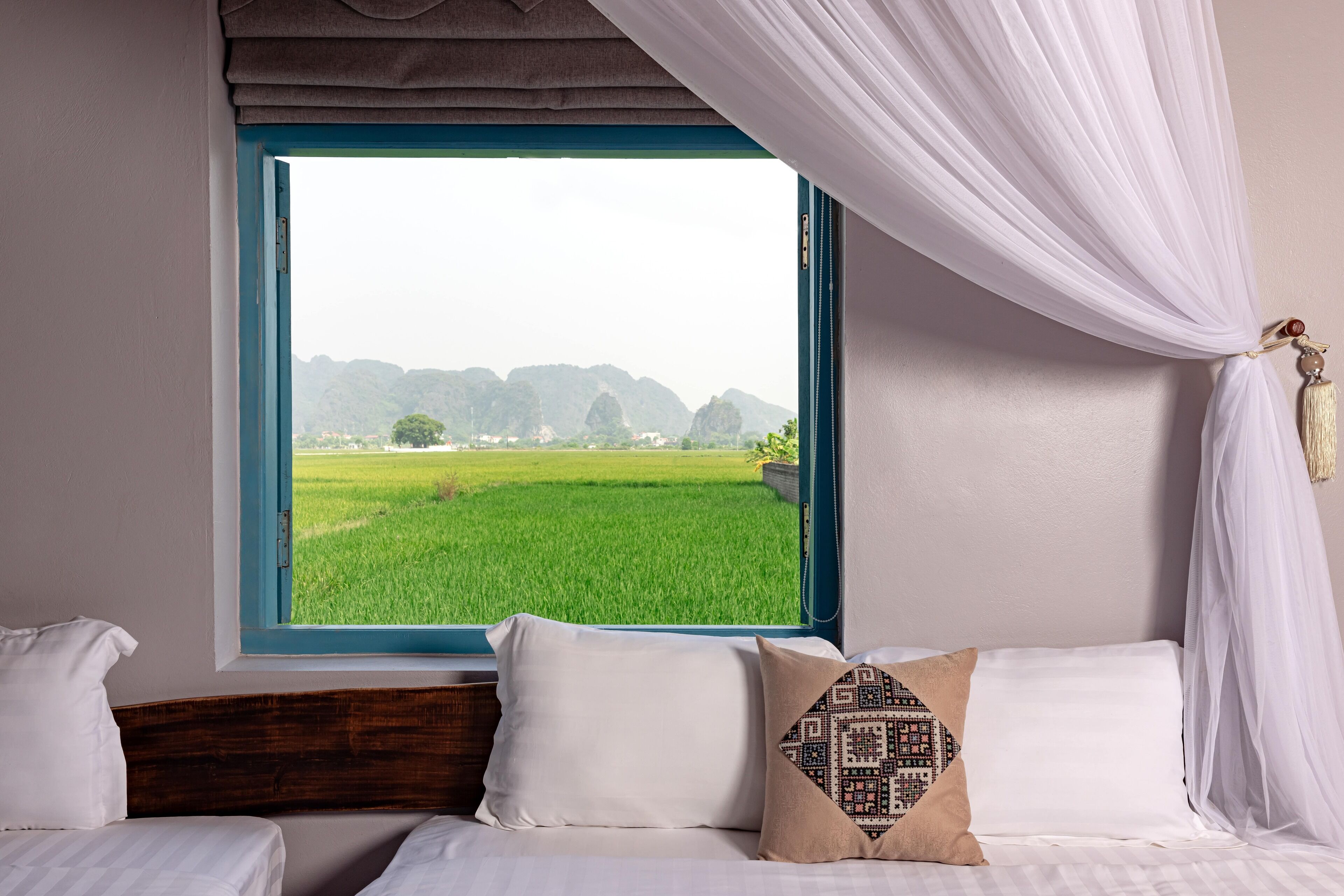 A bedroom with a large window, a bed with white bedding, and a view of a green field and hills.