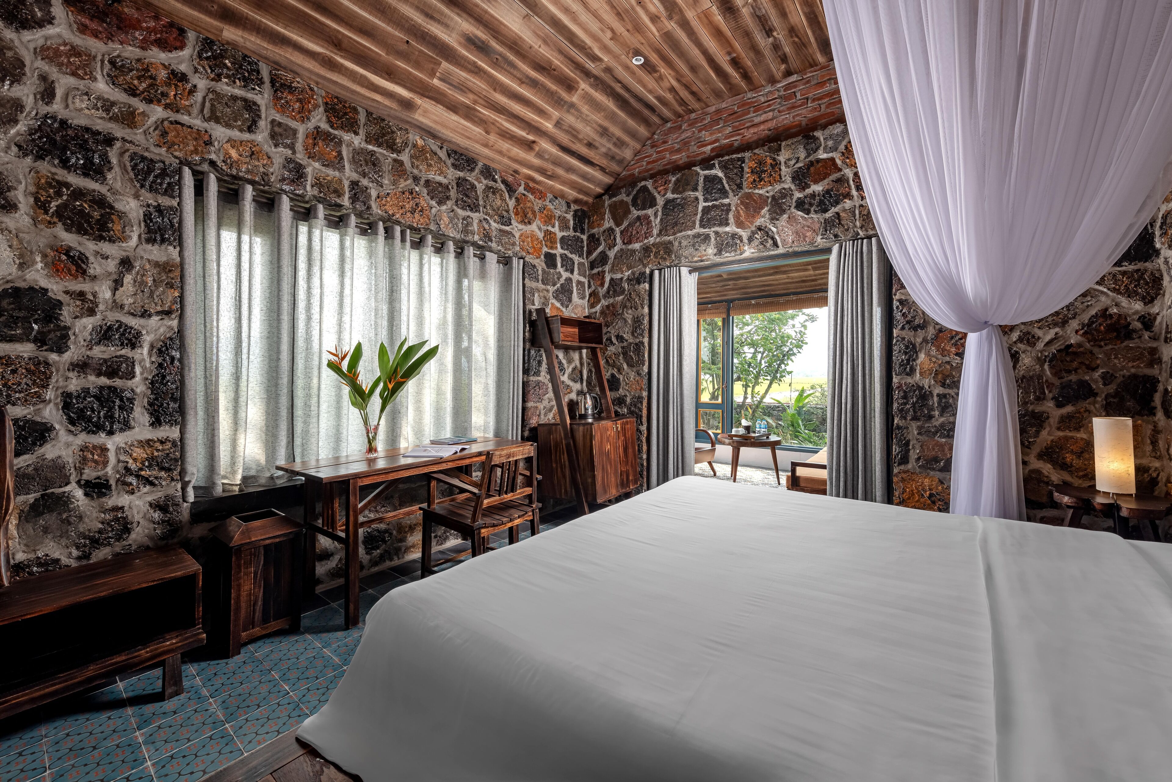 A bedroom with a stone wall, wooden ceiling, a large bed, a desk with a chair, and a window with curtains.