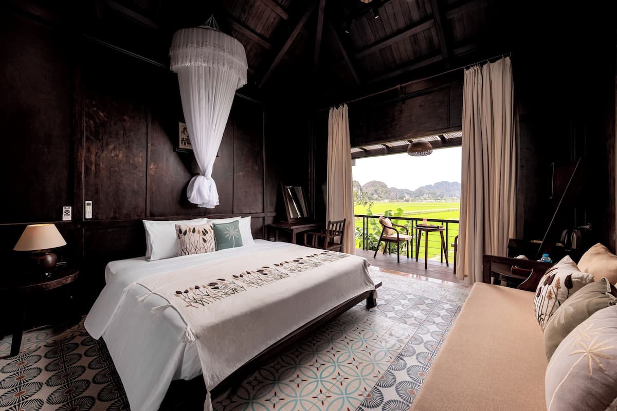 A bedroom with a large bed, a nightstand, a chair, and a view of a green landscape through a balcony.