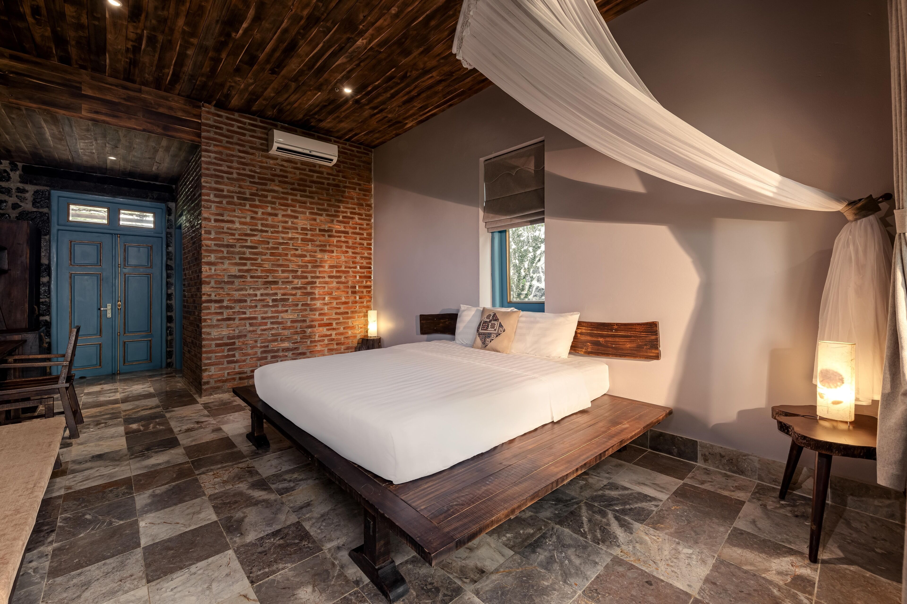 A bedroom with a large bed, a wooden bench, a brick wall, and a blue door.