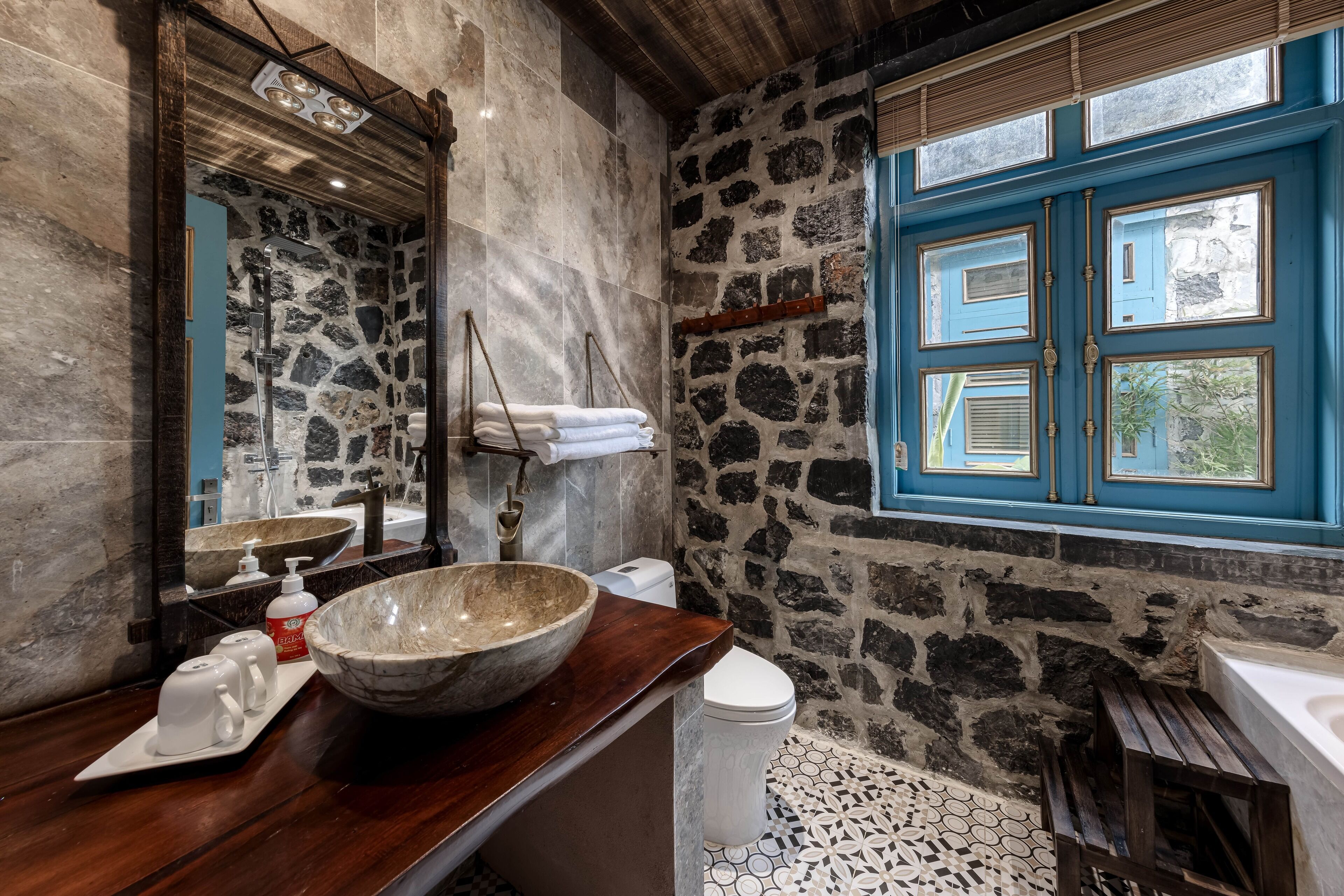 Een badkamer met een stenen muur, een houten wastafelmeubel, een grote wasbak, een wit toilet en een raam met blauwe luiken.