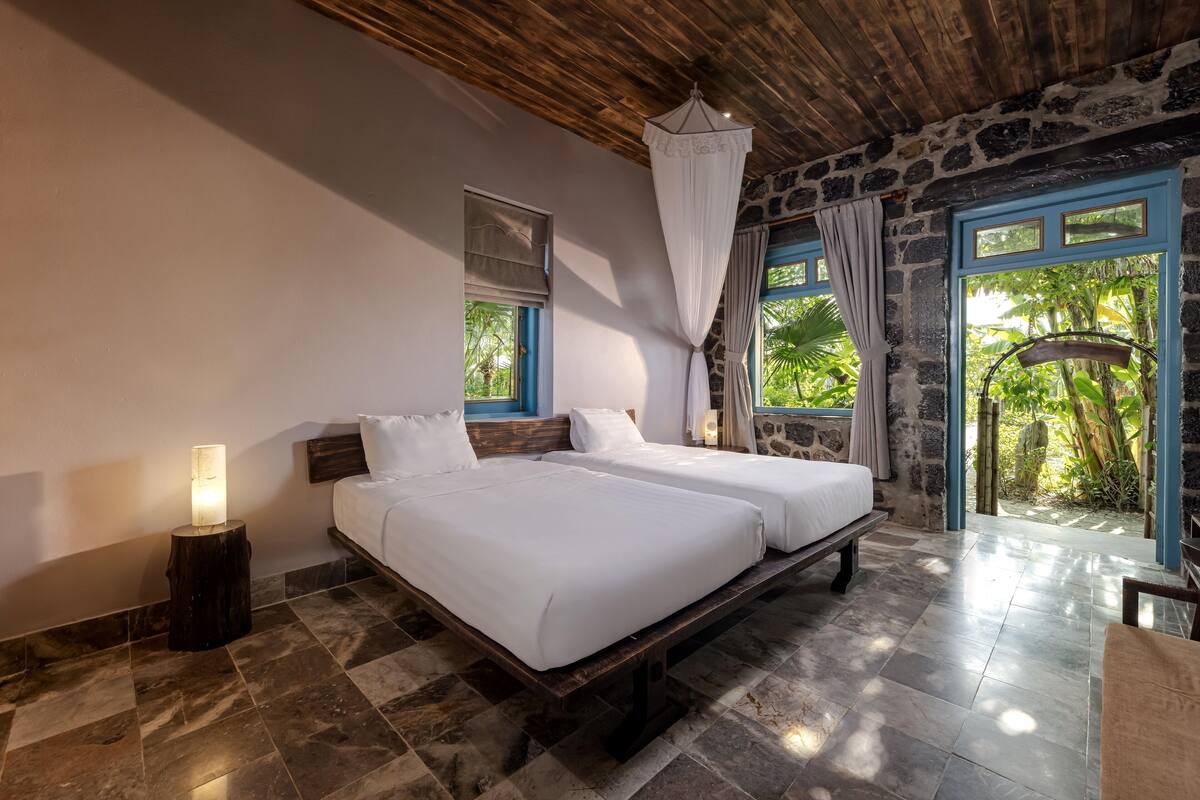 A bedroom with two beds, a stone wall, a wooden ceiling, and a view of greenery.