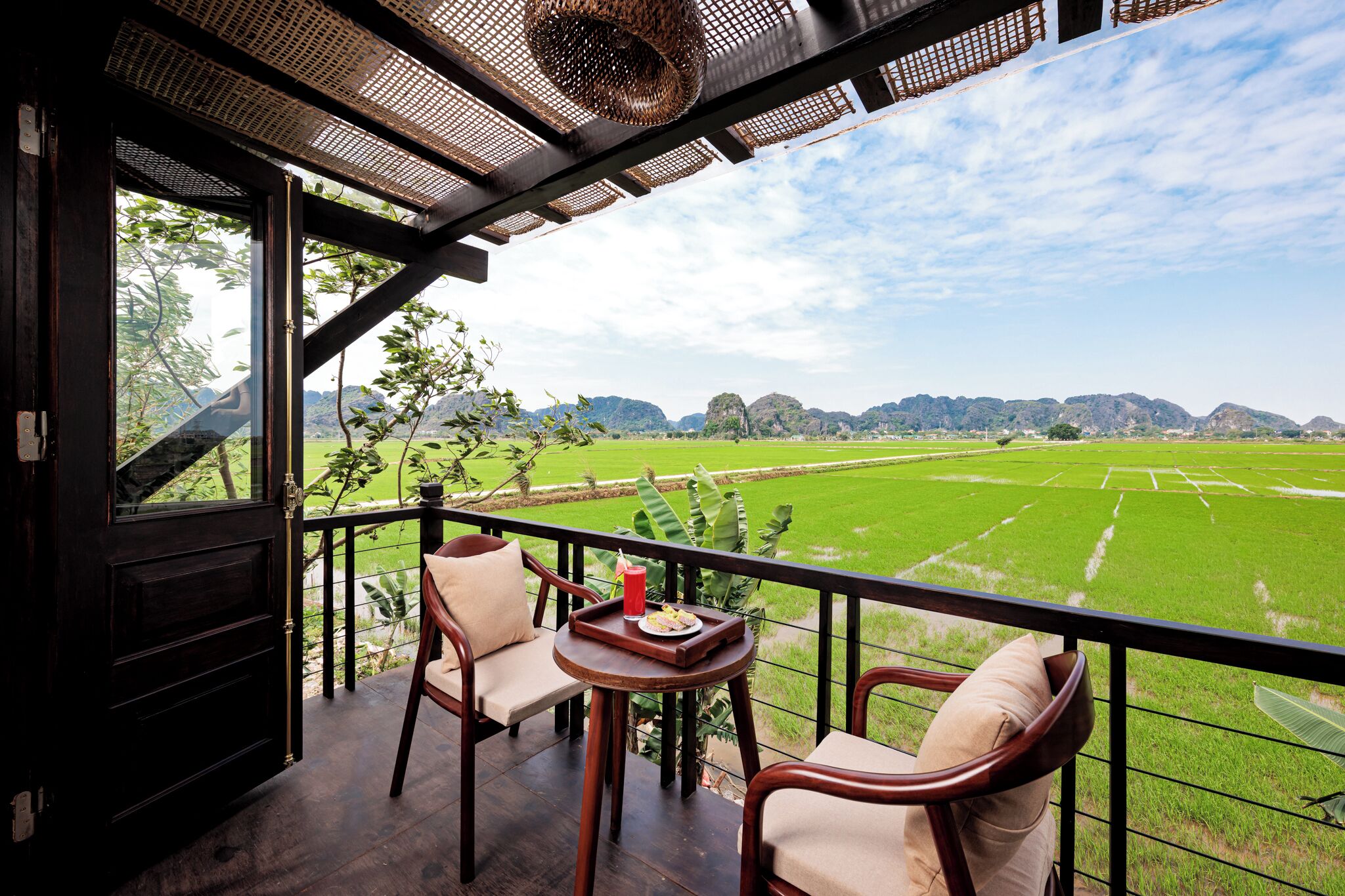 A balcony with two chairs, a small table with a tray, and a view of a green field and mountains.